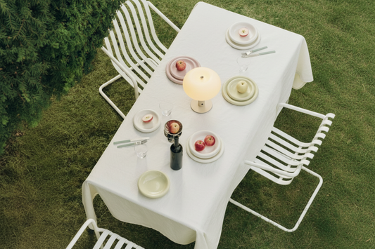 Cream cordless table lamp with warm glowing opal glass shade styled on an outdoor white tablecloth dining table with pastel ceramic plates, apples, and wine bottle in a garden setting