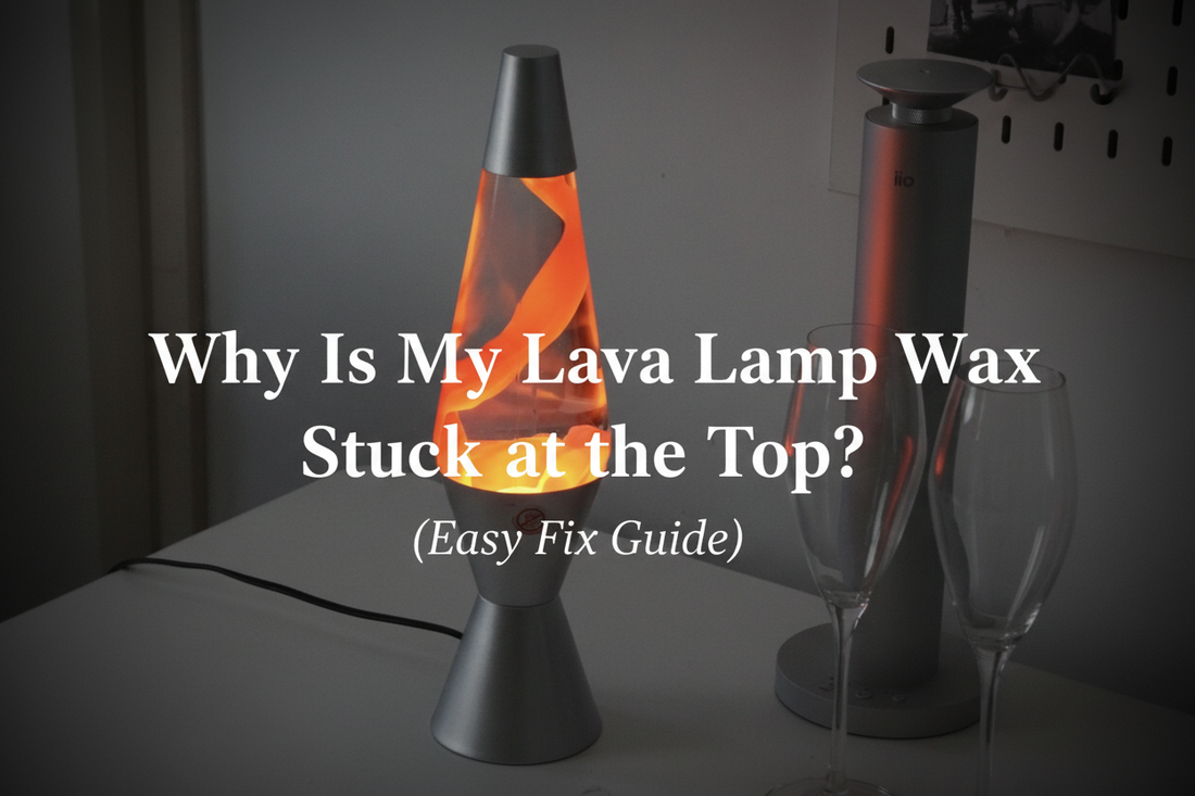 A silver lava lamp with glowing orange wax forming large swirling ribbon-like shapes inside, placed on a grey desk next to wine glasses and a speaker in a minimal modern setting.