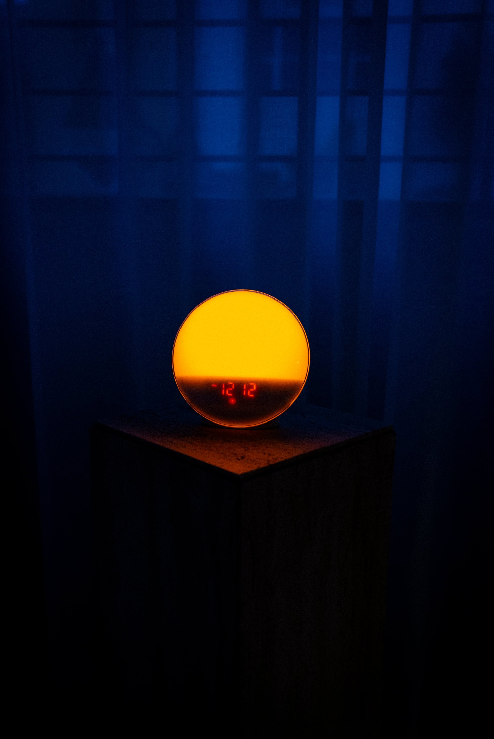 Lumiosa dawn simulator emitting soft light on a pedestal, displaying the time 12:12 in a dimly lit bedroom.