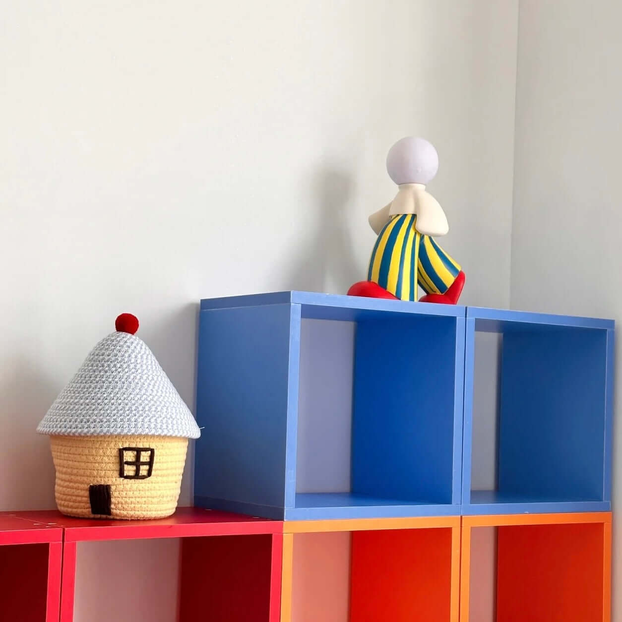 Colorful decor featuring a human-shaped figure lamp and a crocheted house on vibrant cube shelves.