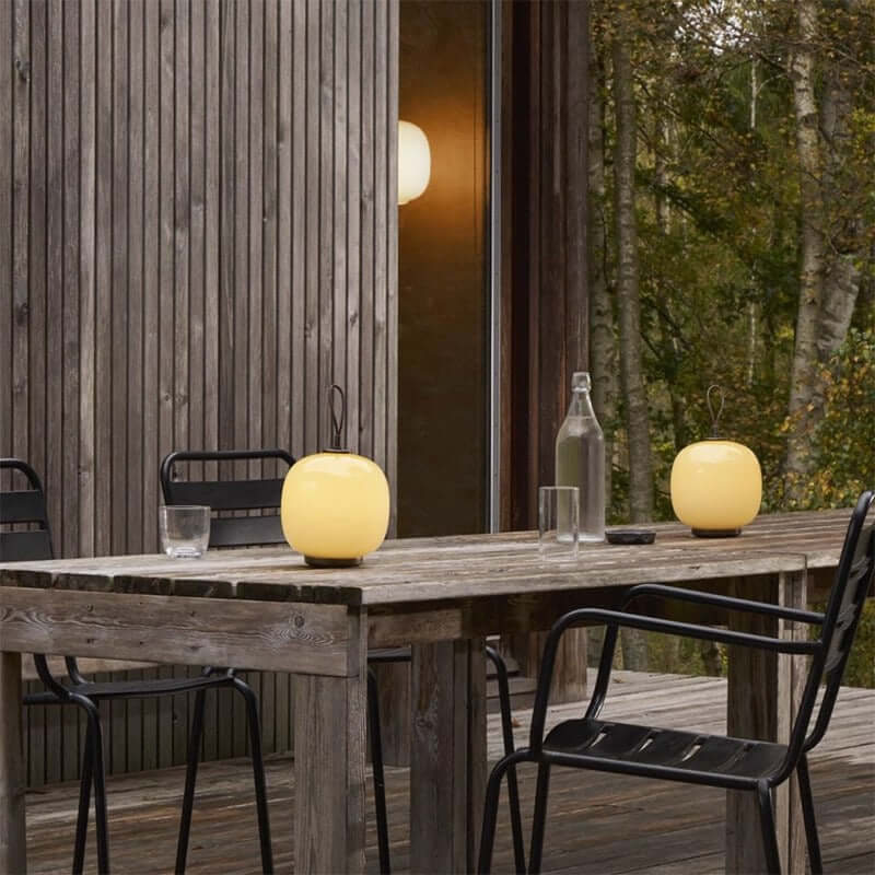 Danish portable table lamps on a wooden outdoor table, creating a cozy ambiance in a nature-inspired setting.