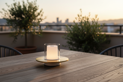 Lumiosa™ Eternal Flame rechargeable candle lamp with brushed brass base glowing warm 2400K light on a rustic oak patio table at golden hour, Mediterranean city skyline in soft bokeh background